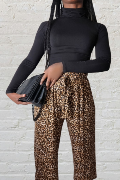 Person wearing a black long-sleeve top and leopard print pants, holding a black clutch against a white brick wall.