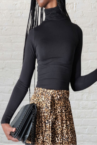 Person wearing a black turtleneck and leopard print skirt holding a black clutch against a light gray brick wall.