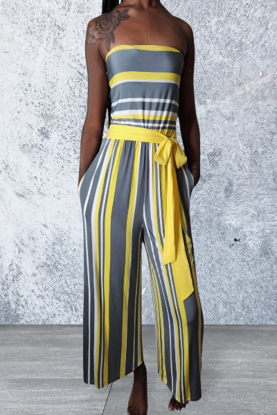 Woman wearing a strapless, yellow, and gray striped jumpsuit against a textured white wall.