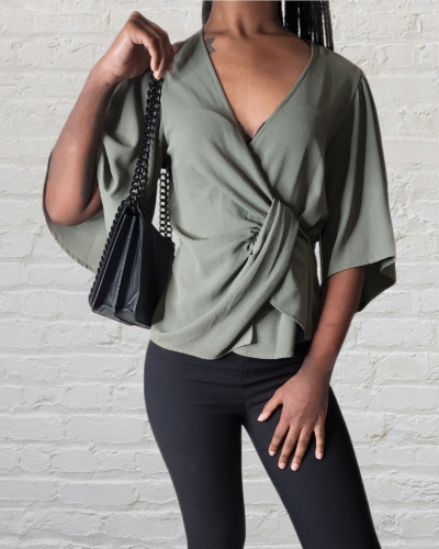 Woman wearing a green top and black pants, holding a black handbag against a white brick wall.