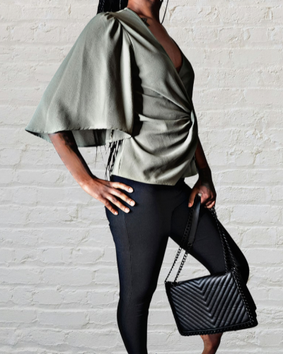 Person wearing a green top and black leggings holding a black clutch against a white brick wall.